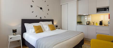 A modern bedroom with a large bed, bedside tables, a clock on the wall, and a kitchenette with a microwave.