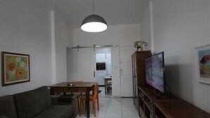 Ein Wohnzimmer mit einer Couch, einem Esstisch mit Stühlen, einem Fernseher und einer Küche im Hintergrund.