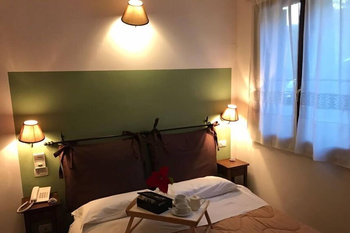 A hotel room with a bed, bedside lamps, a bedside phone, and a tray table with a teapot and cups.