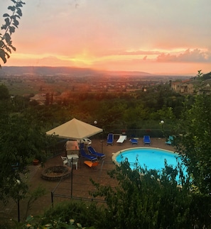 A swimming pool with lounge chairs and a sun umbrella, surrounded by greenery and overlooking a distant landscape.