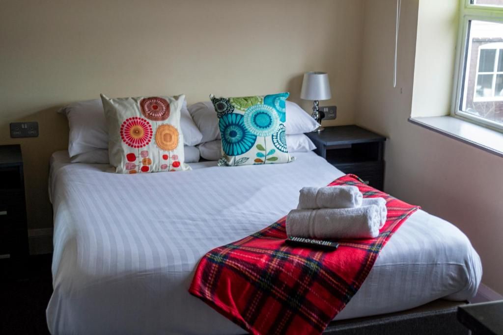 A hotel room with a bed, pillows, a red plaid blanket, and a nightstand with a lamp.