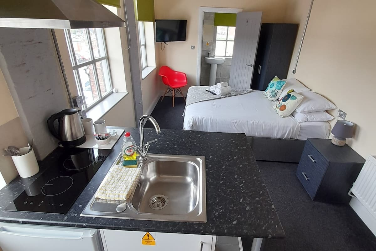 A compact hotel room with a kitchenette, a bed with pillows, a red chair, and a window.