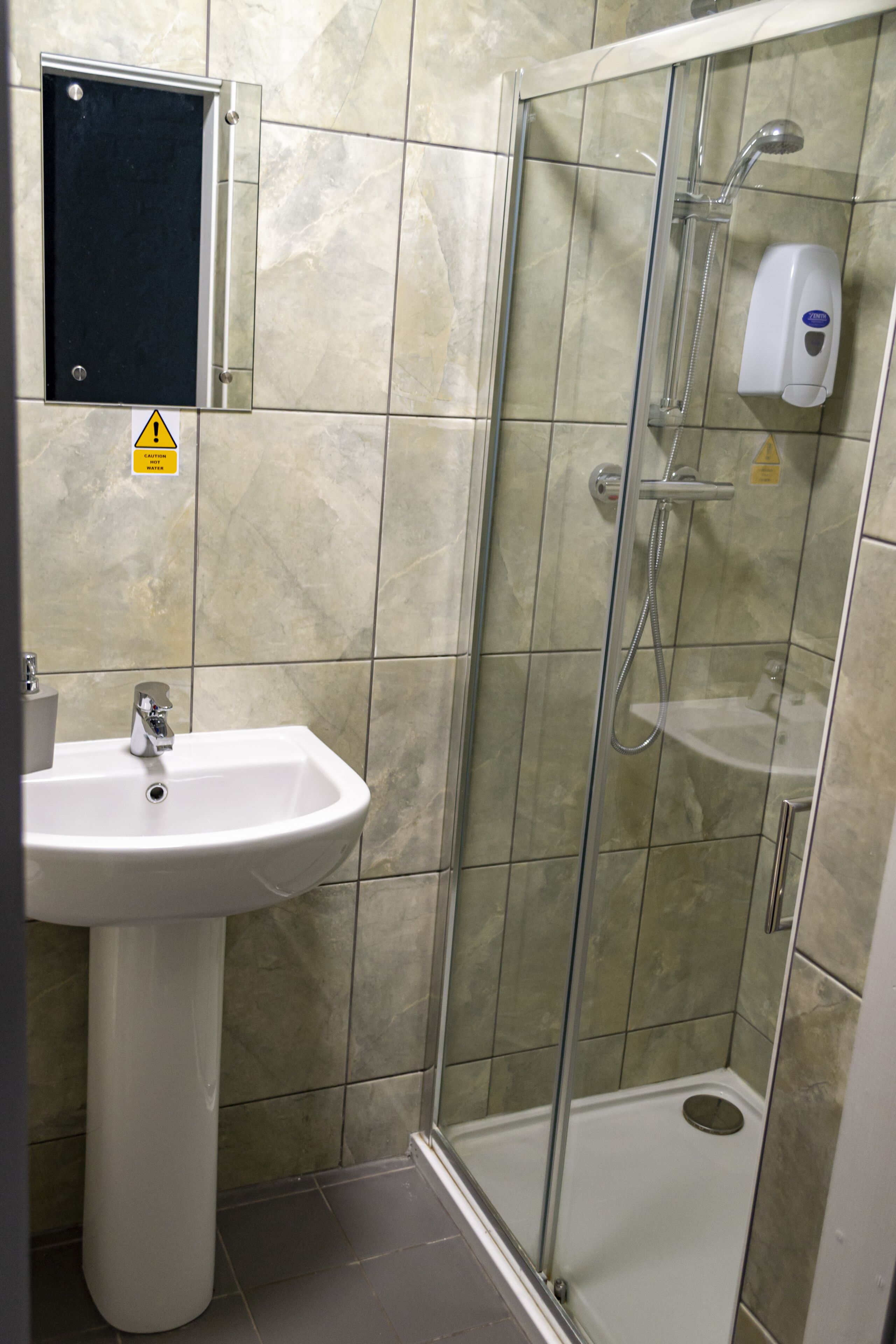 A bathroom with a glass-enclosed shower, a pedestal sink, and a hand soap dispenser.
