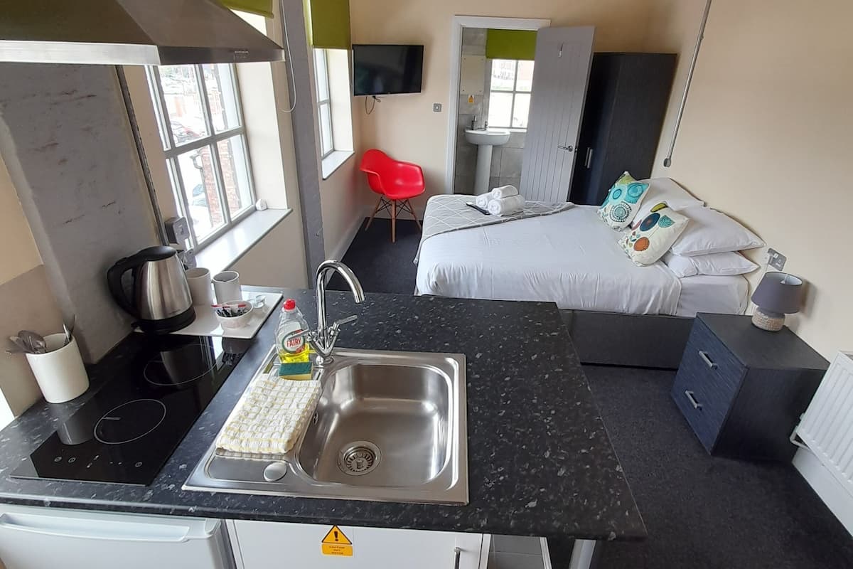 A compact hotel room with a kitchenette, a bed with pillows, a red chair, and a bathroom with a sink and mirror.