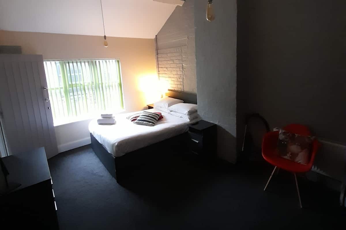 A bedroom with a bed, a desk, a chair, and a window with blinds.
