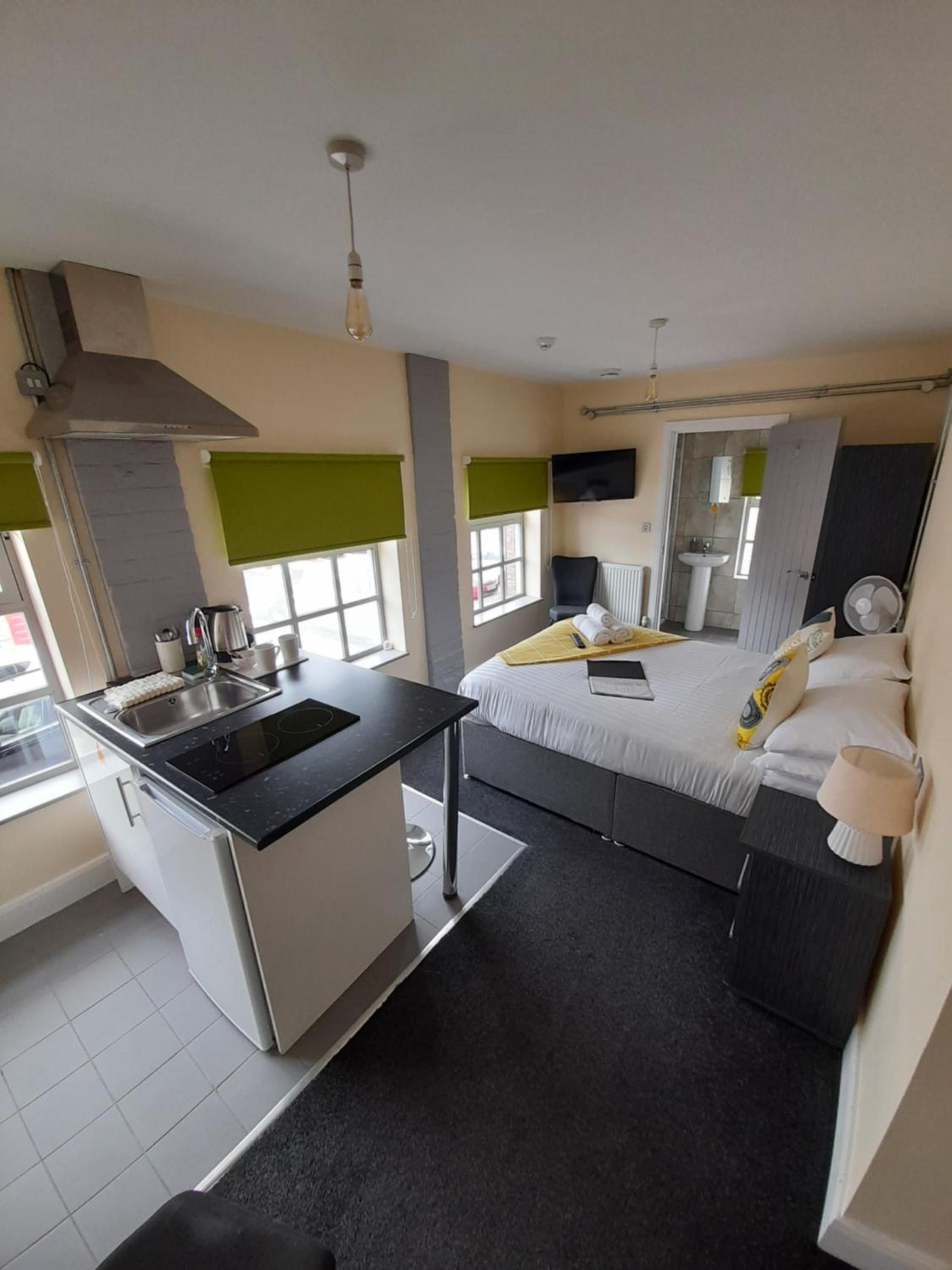 A compact hotel room with a kitchenette, a bed with pillows, a bedside table, a TV mounted on the wall, and a window with blinds.