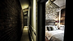 A narrow hallway with a brick wall on one side and a bed with a patterned bedspread and pillows on the other. - The Cow Hollow Hotel (Manchester)
