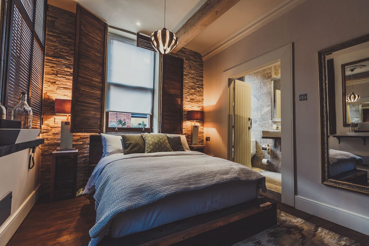 A bedroom with a large bed, wooden walls, a stone accent wall, a window, a mirror, and a nightstand with a lamp.