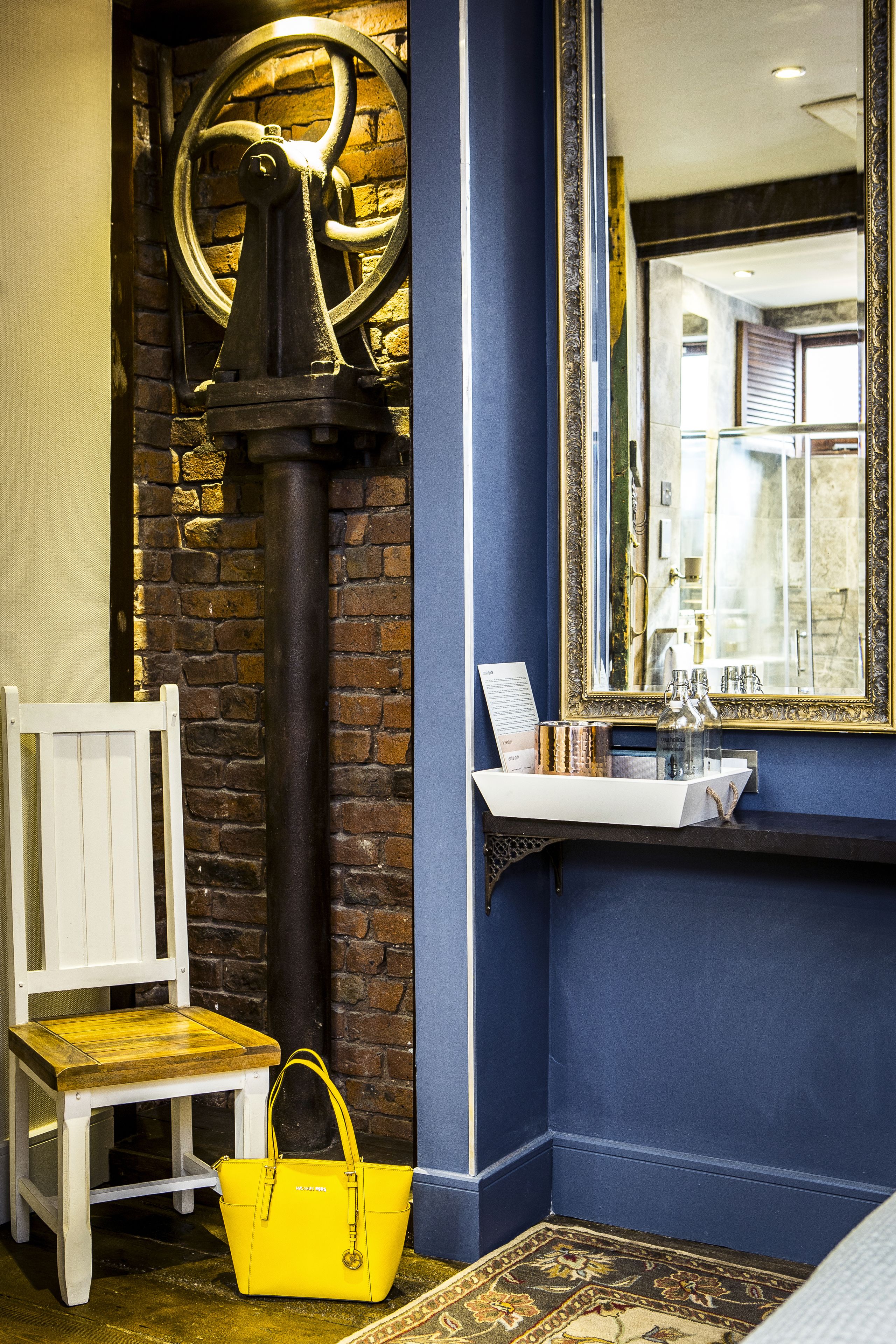A room with a brick wall, a white chair, a yellow handbag, a black pipe, and a mirror with a gold frame.
