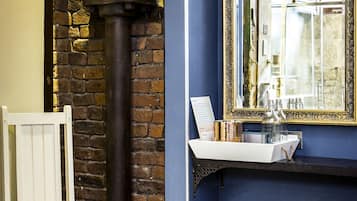 A room with a brick wall, a white chair, a yellow handbag, a black pipe, and a mirror with a gold frame.