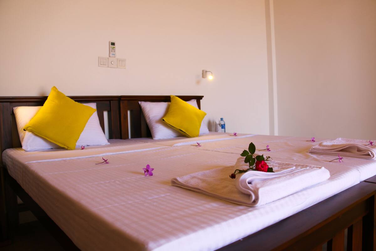 A hotel room with a double bed, two pillows, a bedside table with a towel and a flower, and a wall-mounted light.