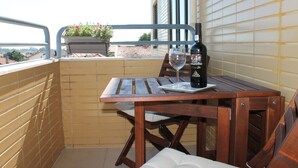 Une table en bois avec une bouteille de vin et un verre, sur un balcon dont le mur est recouvert de carreaux et où se trouve un pot de fleurs.
