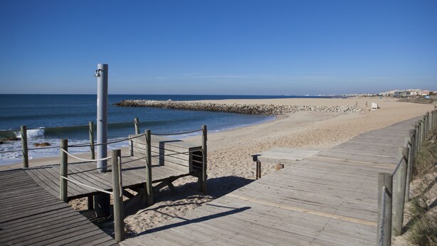 Une promenade en bois mène à une plage de sable, bordée d’un brise-lames rocheux, sous un ciel bleu et dégagé.