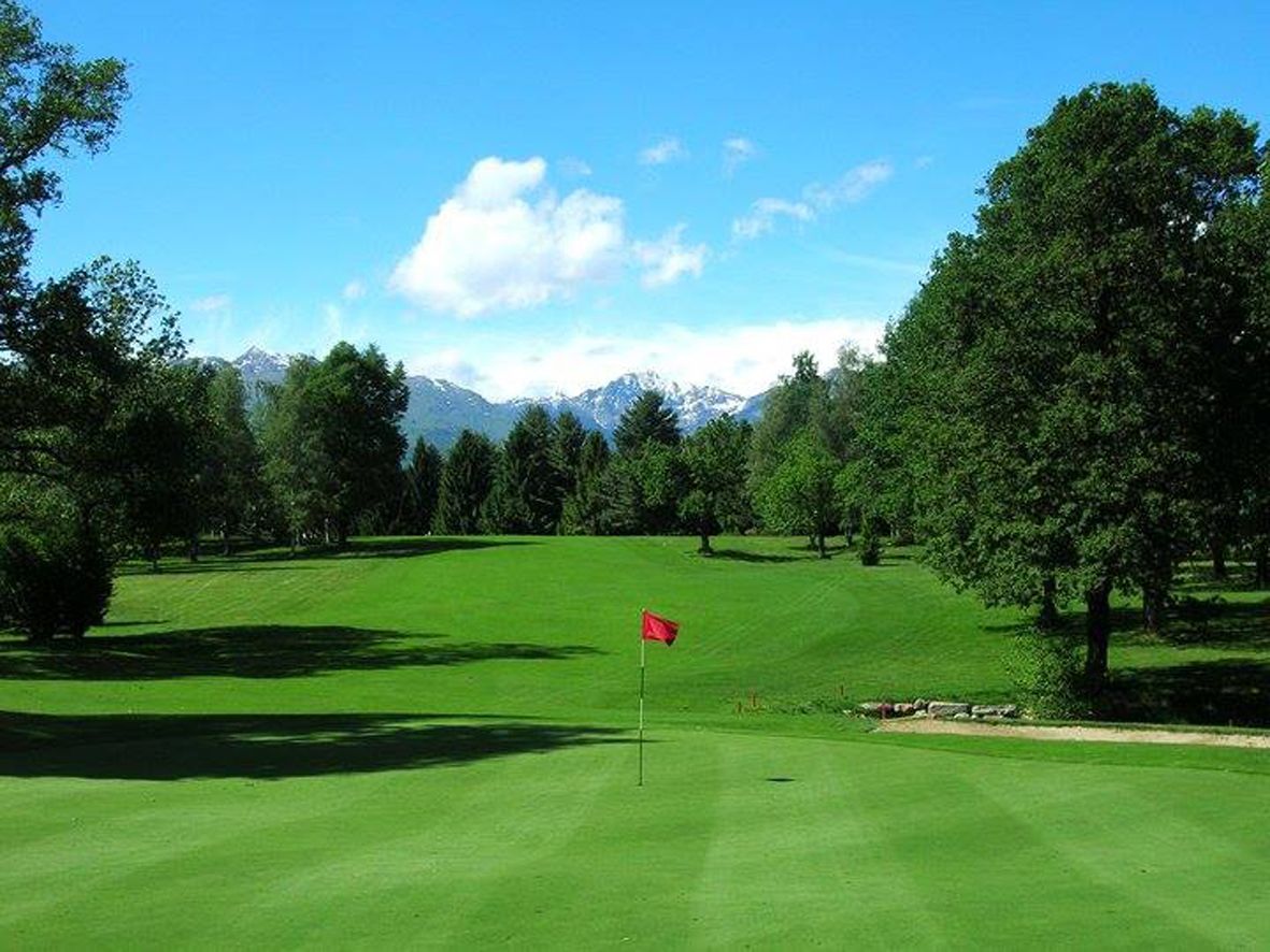 Un parcours de golf avec un drapeau rouge sur le green, entouré d’arbres et avec des montagnes en arrière-plan.