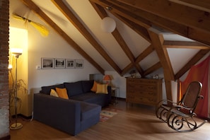 A cozy loft-style room with a dark blue sofa, a rocking chair, wooden beams, a brick wall, and a floor lamp.