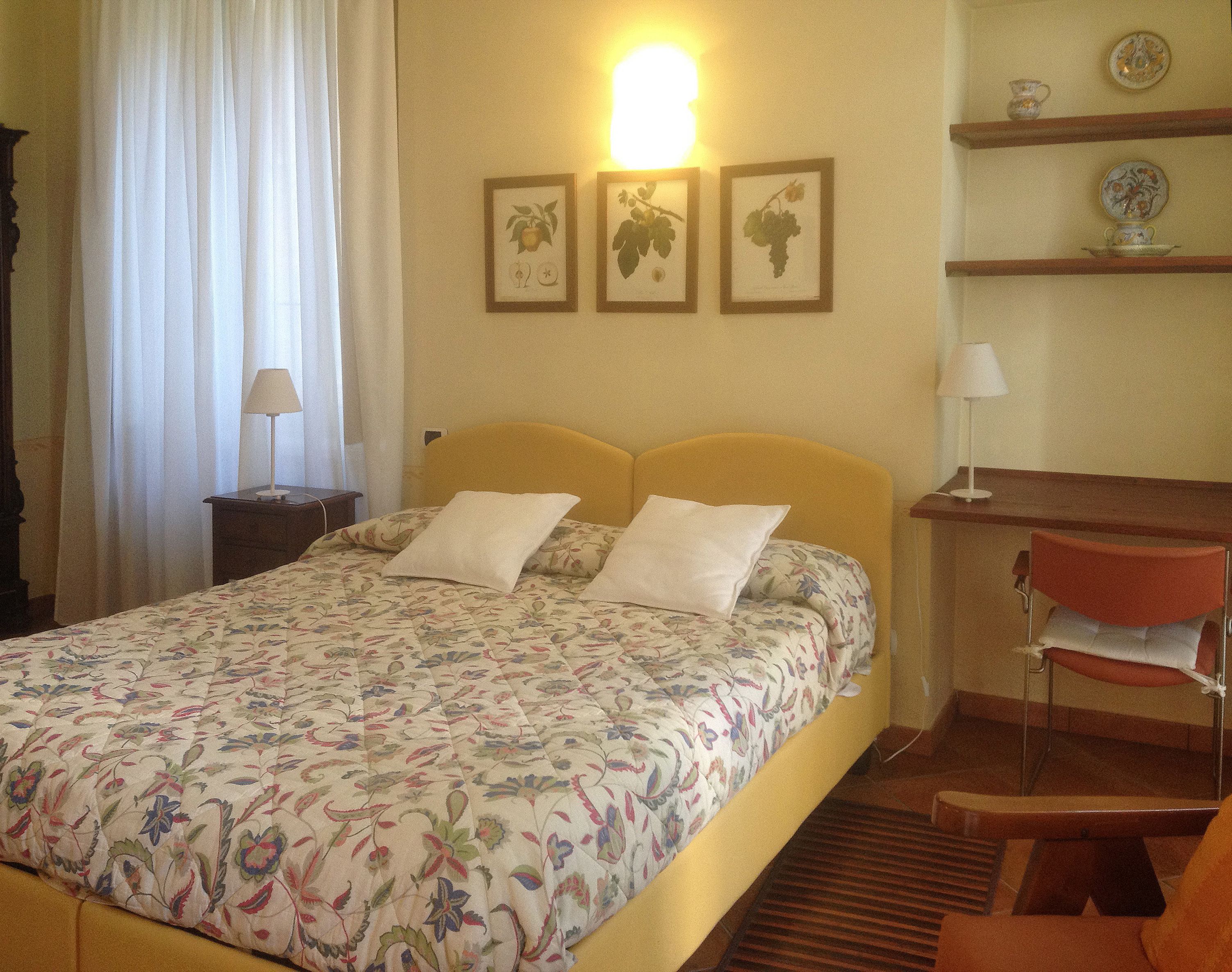 Quarto de hotel com uma cama, uma escrivaninha, uma cadeira e uma prateleira com objetos decorativos.