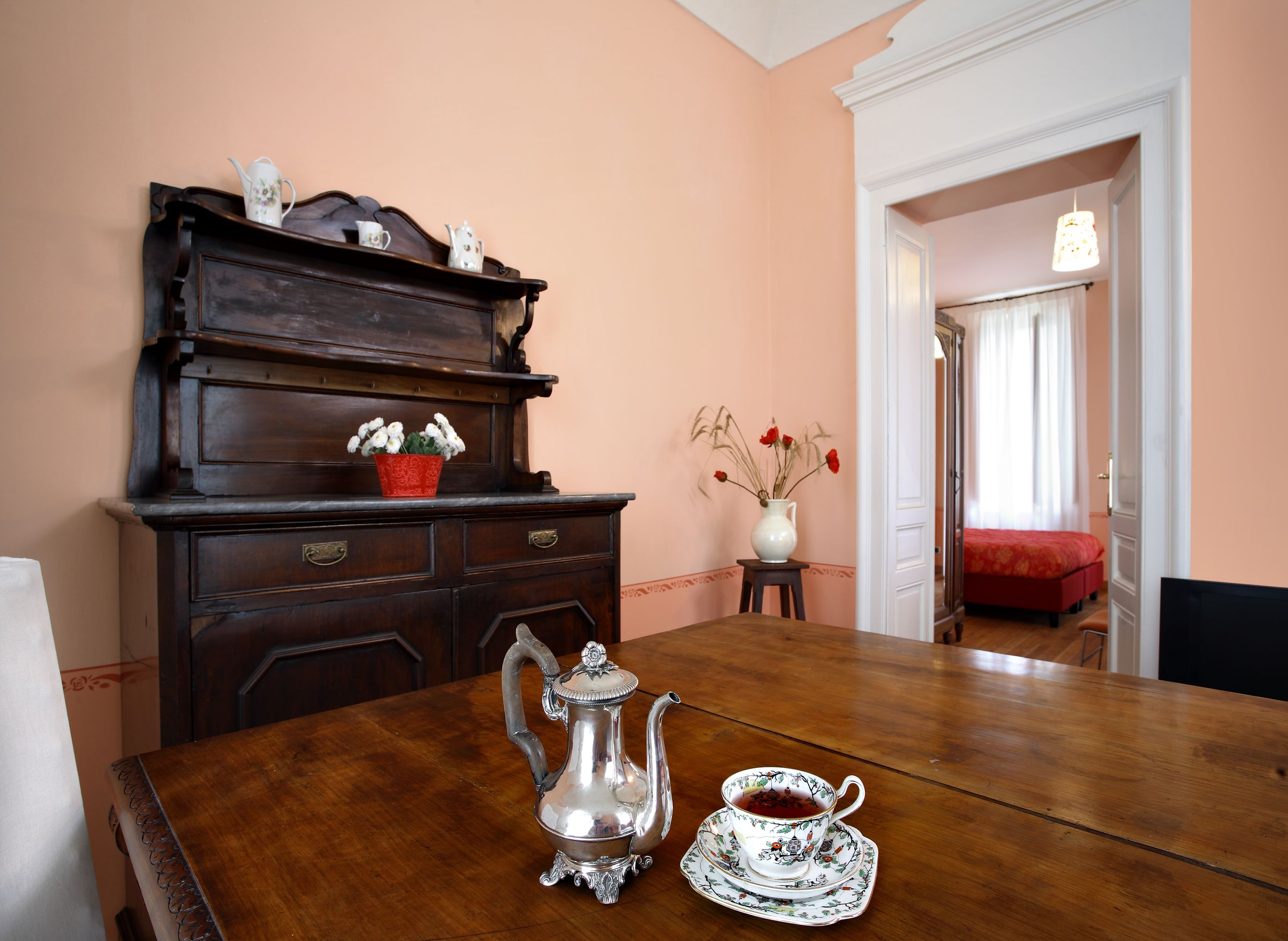 Una mesa de comedor de madera con una tetera de plata y una taza, un florero rojo y un aparador de madera con objetos decorativos.