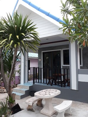 A covered outdoor seating area with a table and benches, surrounded by potted plants and a palm tree.