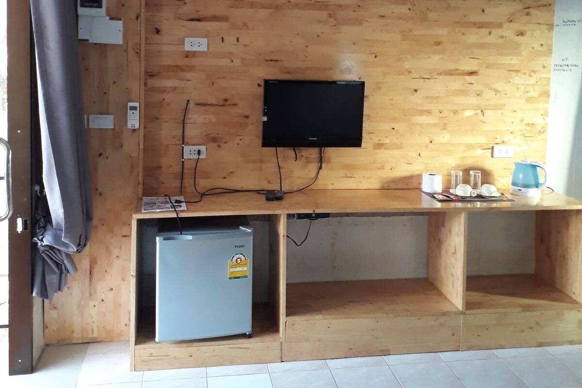 A room with a wooden wall, a flat-screen TV mounted above a wooden desk, a small refrigerator, and a kettle on the desk.