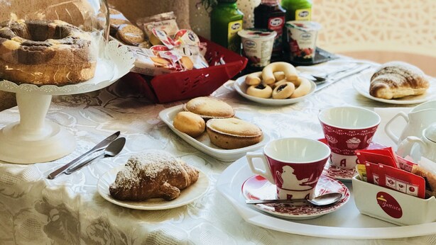 A table set with pastries, a glass dome cake, and a vase of purple flowers. - Villa Aurora B&B (Bari)