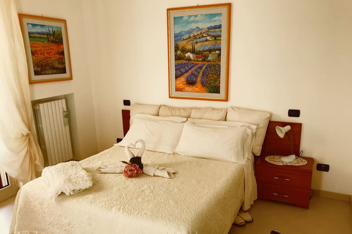 Cama bem arrumada com arranjo de flores, duas pinturas na parede, abajur e mesa de cabeceira vermelha.
