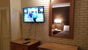 A hotel room with a television mounted on the wall, a bedside table with a lamp, and a mirror reflecting the room's interior. - Americas Premier Inn (Sweetwater)