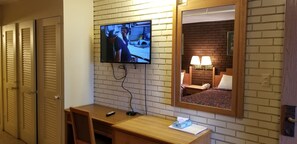 A hotel room with a TV mounted on a brick wall, a wooden desk, a bedside table with a lamp, and a mirror reflecting the room. - Americas Premier Inn (Sweetwater)