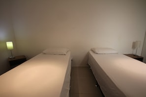 Dos camas individuales con sábanas blancas en una habitación con paredes lisas y piso de baldosas.