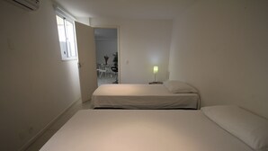 Een kleine, zwak verlichte kamer met twee bedden, een nachtkastje met een lamp en een raam met gesloten jaloezieën.