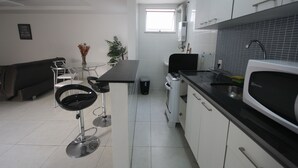 Een moderne keuken met witte kastjes, een zwart aanrecht en een roestvrijstalen spoelbak. Er is een eethoek met een glazen tafel en twee zwarte barkrukken. Een klein raam zorgt voor natuurlijk licht.