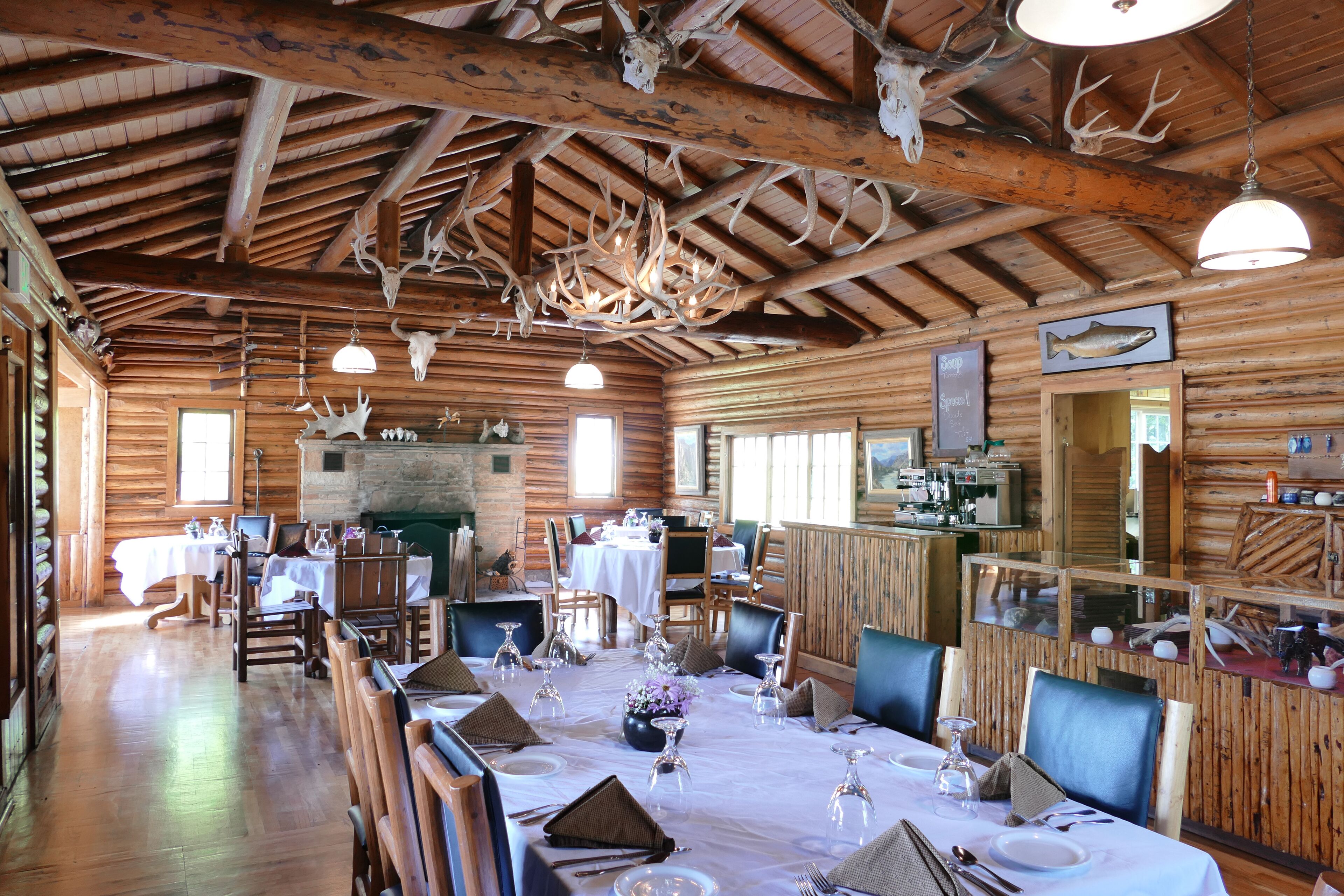 A log cabin restaurant with a rustic interior, wooden beams, antler chandeliers, and a dining area set for a meal.