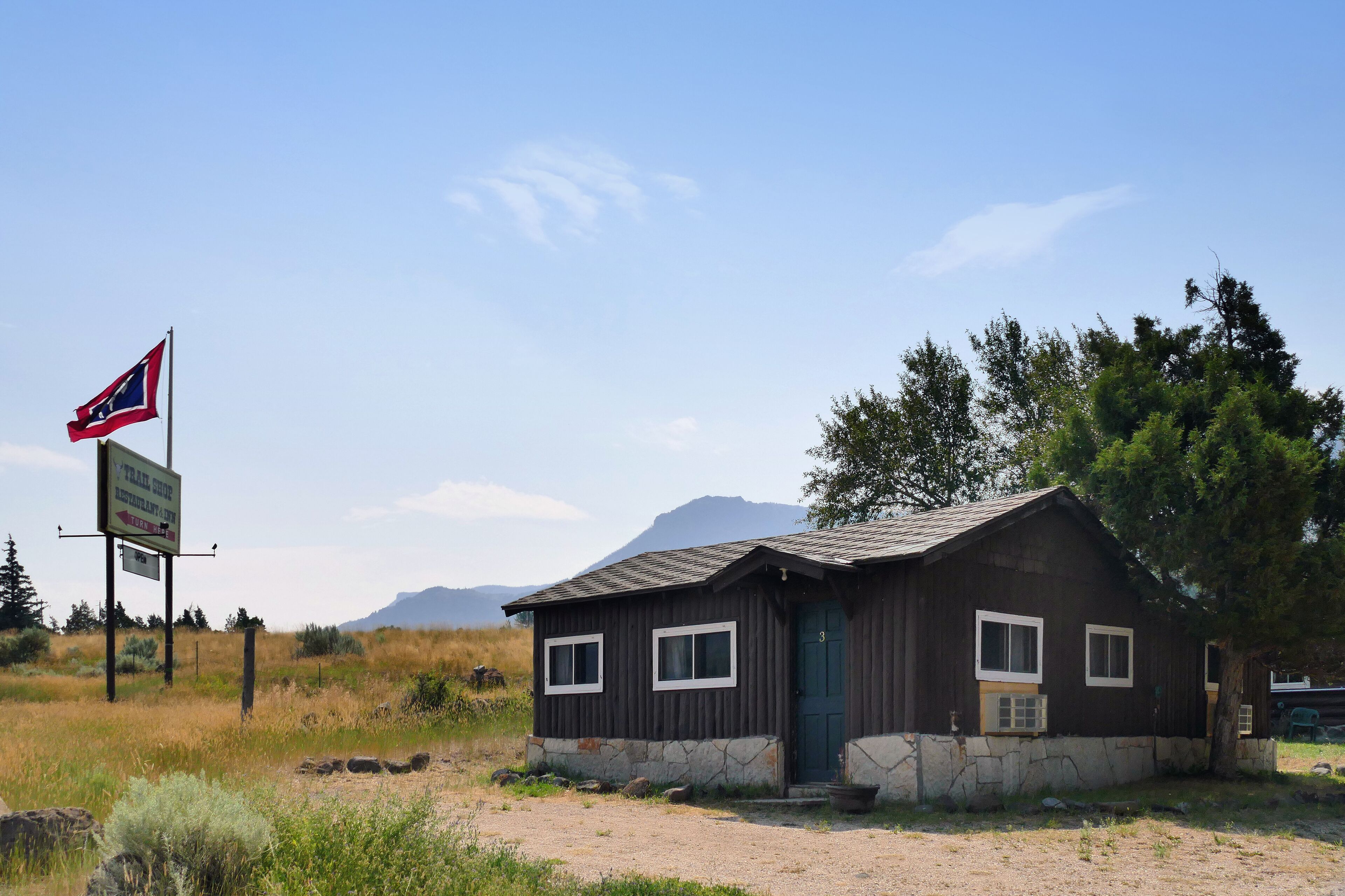 A rustic cabin with a stone foundation and a dark wooden exterior, surrounded by a natural landscape with a mountain in the background.