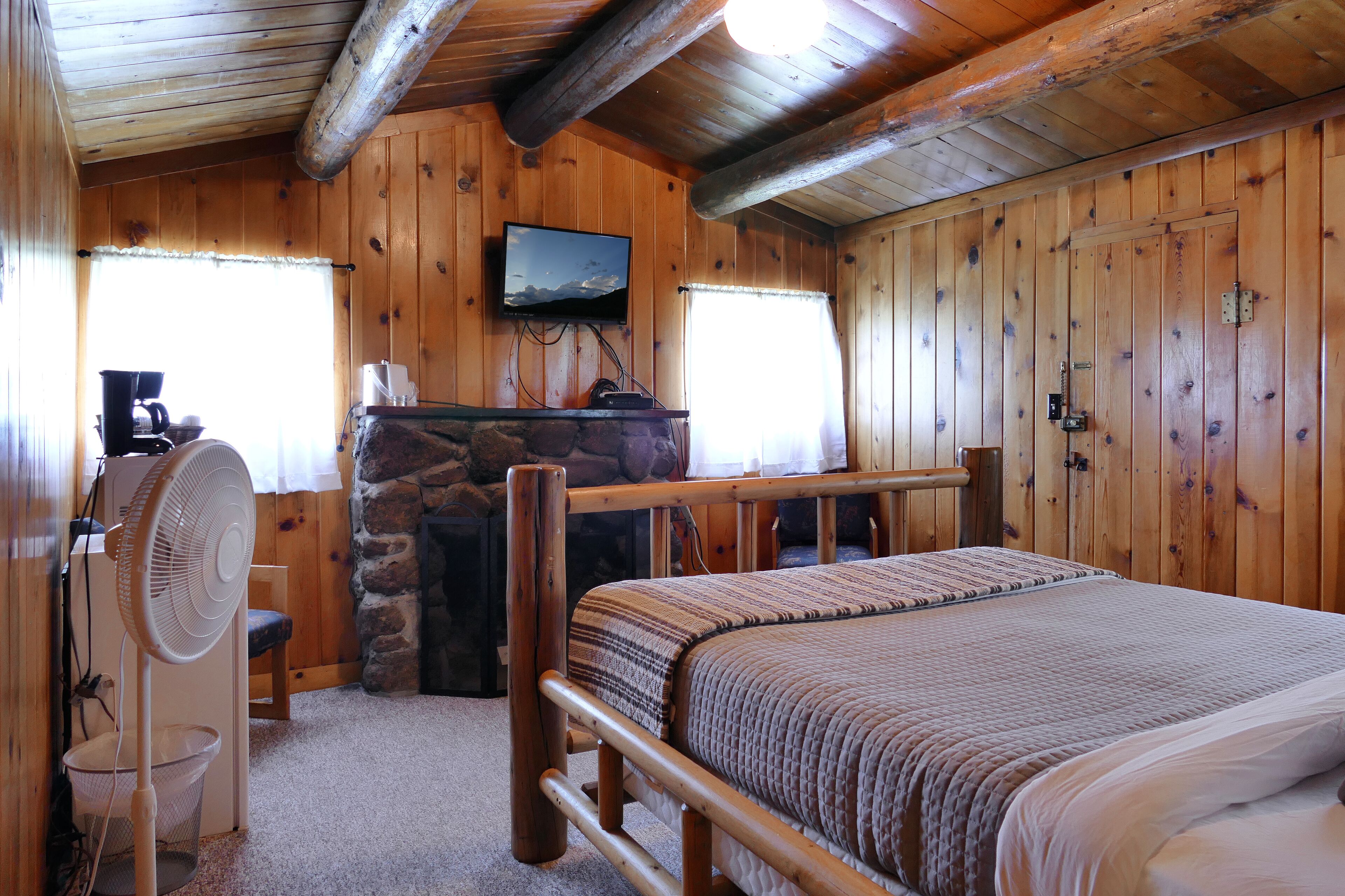 A wooden cabin room with a bed, a stone fireplace, a television, and a fan.