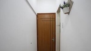 Un pasillo estrecho con una puerta de madera al final, paredes blancas y un estante pequeño con objetos en la pared derecha.