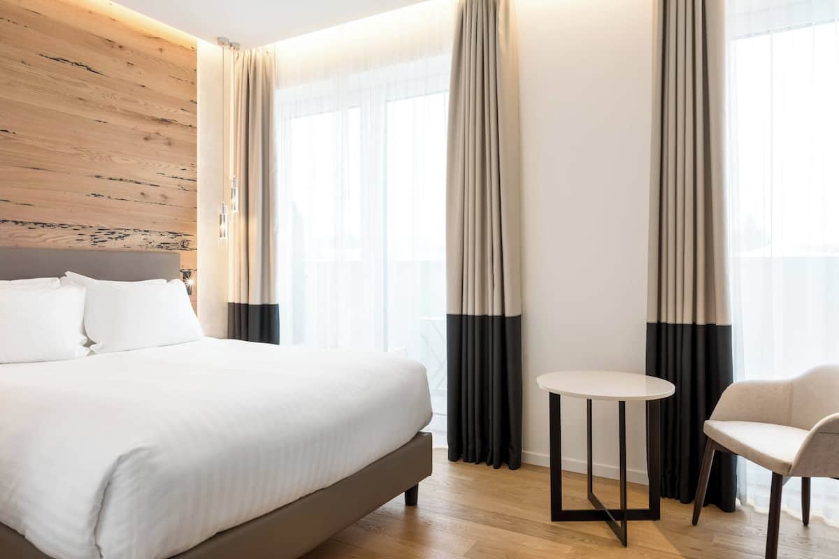 A hotel room with a large bed, a small round table, a chair, and a view of the city through large windows.
