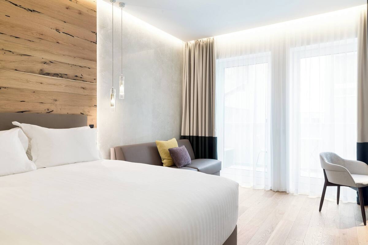 A modern hotel room with a large bed, a sofa, a chair, and a window with curtains.