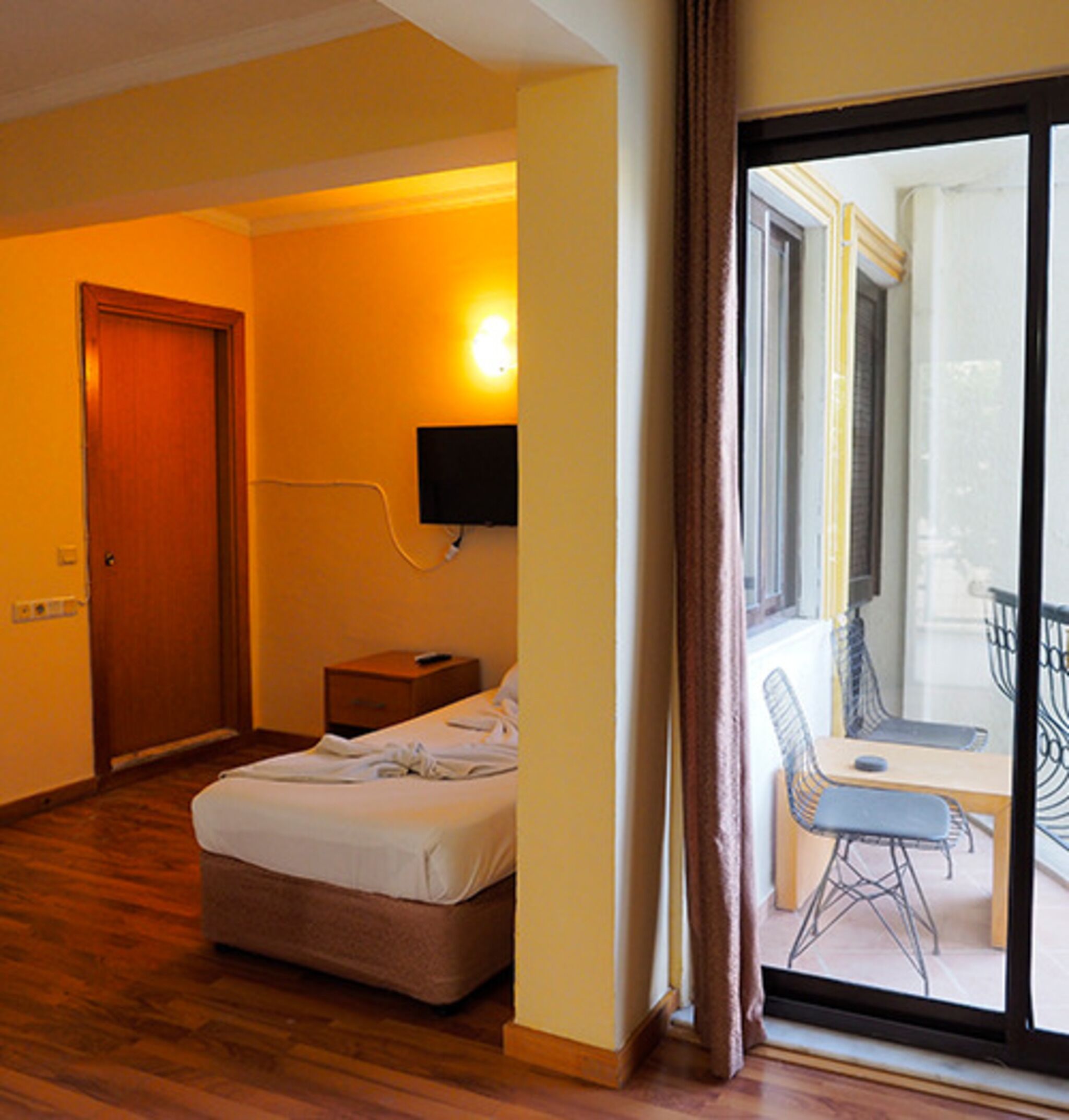 A hotel room with a bed, a bedside table, a TV, and a view of an outdoor balcony.