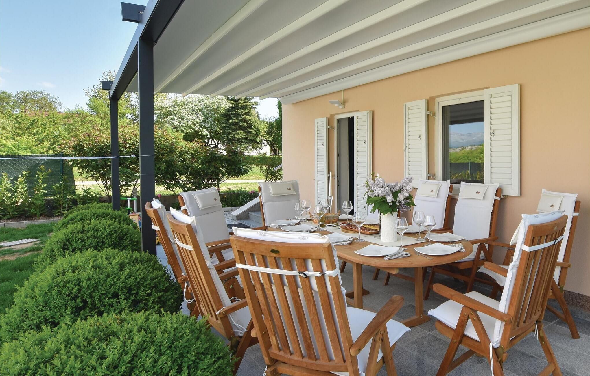 A covered outdoor dining area with a round table set for six, surrounded by wooden chairs with white cushions.