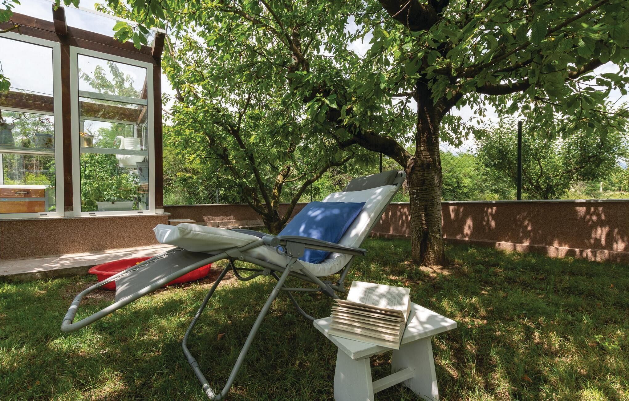 A sun lounger with a book on a stand, a red chair, and a glass window overlooking a garden.