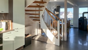 Une cuisine moderne avec des meubles blancs, un escalier en bois et une vue sur une maison, visible à travers une grande fenêtre.