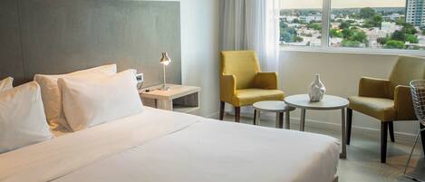 Habitación de hotel con una cama grande, dos sillones, una mesita y vistas a la ciudad a través de un amplio ventanal.