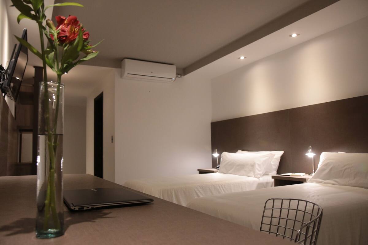 Une chambre d’hôtel avec deux lits, un ordinateur portable sur la table, un vase avec des fleurs et un appareil de climatisation fixé au mur.