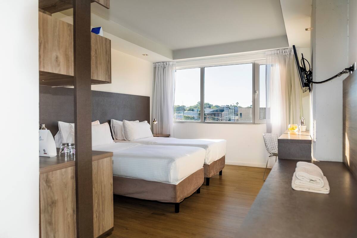 Une chambre d’hôtel avec deux lits, un bureau en bois, une petite table avec une lampe et une grande fenêtre donnant sur des immeubles.