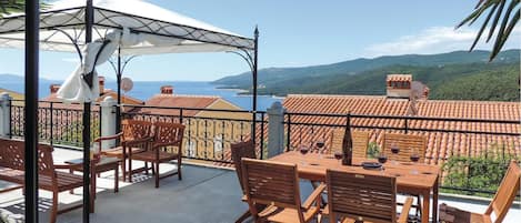 A terrace with wooden furniture, a white umbrella, and a view of the sea and hills.