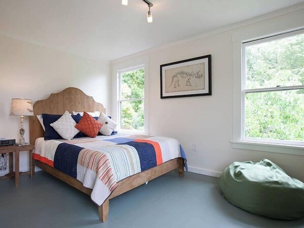 A bedroom with a bed, a bedside table, a lamp, a framed picture, and large windows.