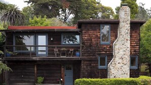 Una casa de madera de dos pisos con chimenea de piedra, un balcón con sillas y un seto bien cuidado.