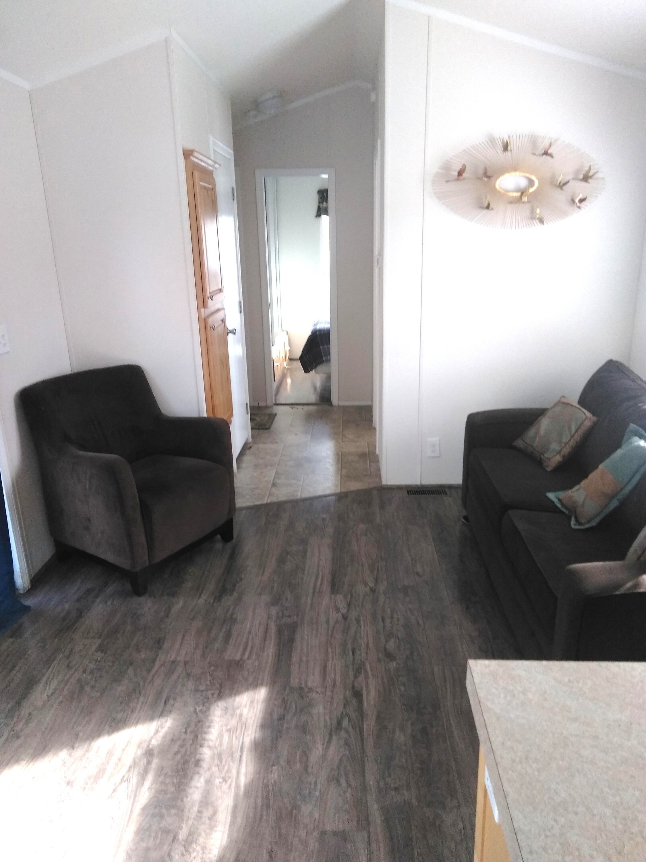 A living room with dark wood flooring, a black sofa with cushions, and a black armchair. A hallway leads to another room with a bed and a window.