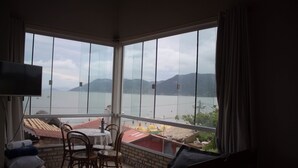A room with a balcony overlooking a body of water and mountains. There is a table with chairs, a TV, and a sofa. - Pousada Estrelas no Mar (Florianópolis)