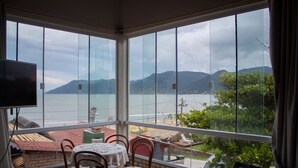 A room with a view of a body of water and mountains. It includes a table with chairs, a television, and a window with curtains. - Pousada Estrelas no Mar (Florianópolis)