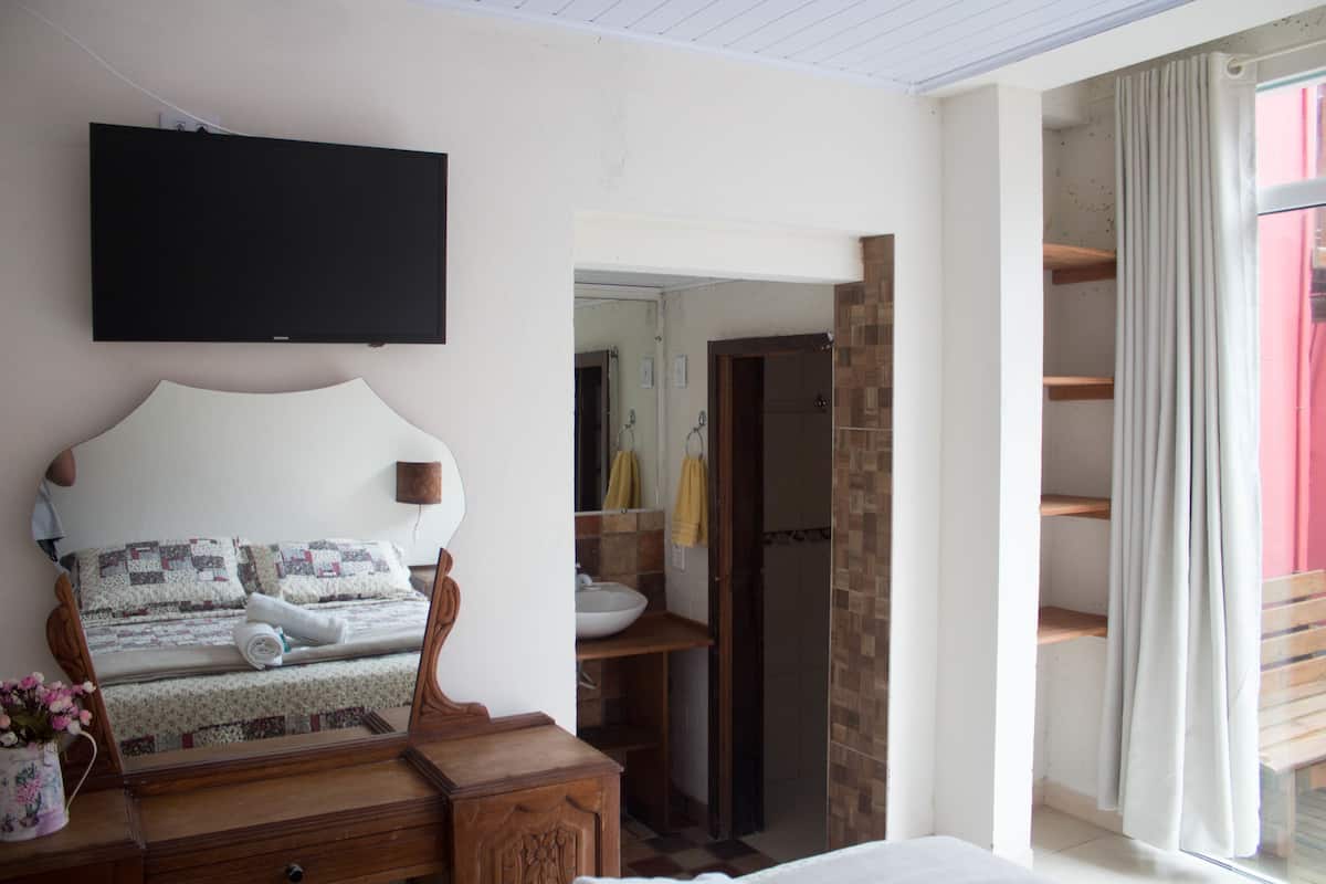 Un dormitorio con cama, mesita de noche, espejo, televisor montado en la pared, baño visible a través de una puerta abierta y ventana con cortinas.